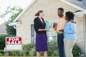 A real estate agent standing in front of a house with a FOR SALE sign in the yard, talking with an African American couple who could be the homeowers, or potential buyers. The agent is wearing a black jacket and purple dress, carrying a blue binder.